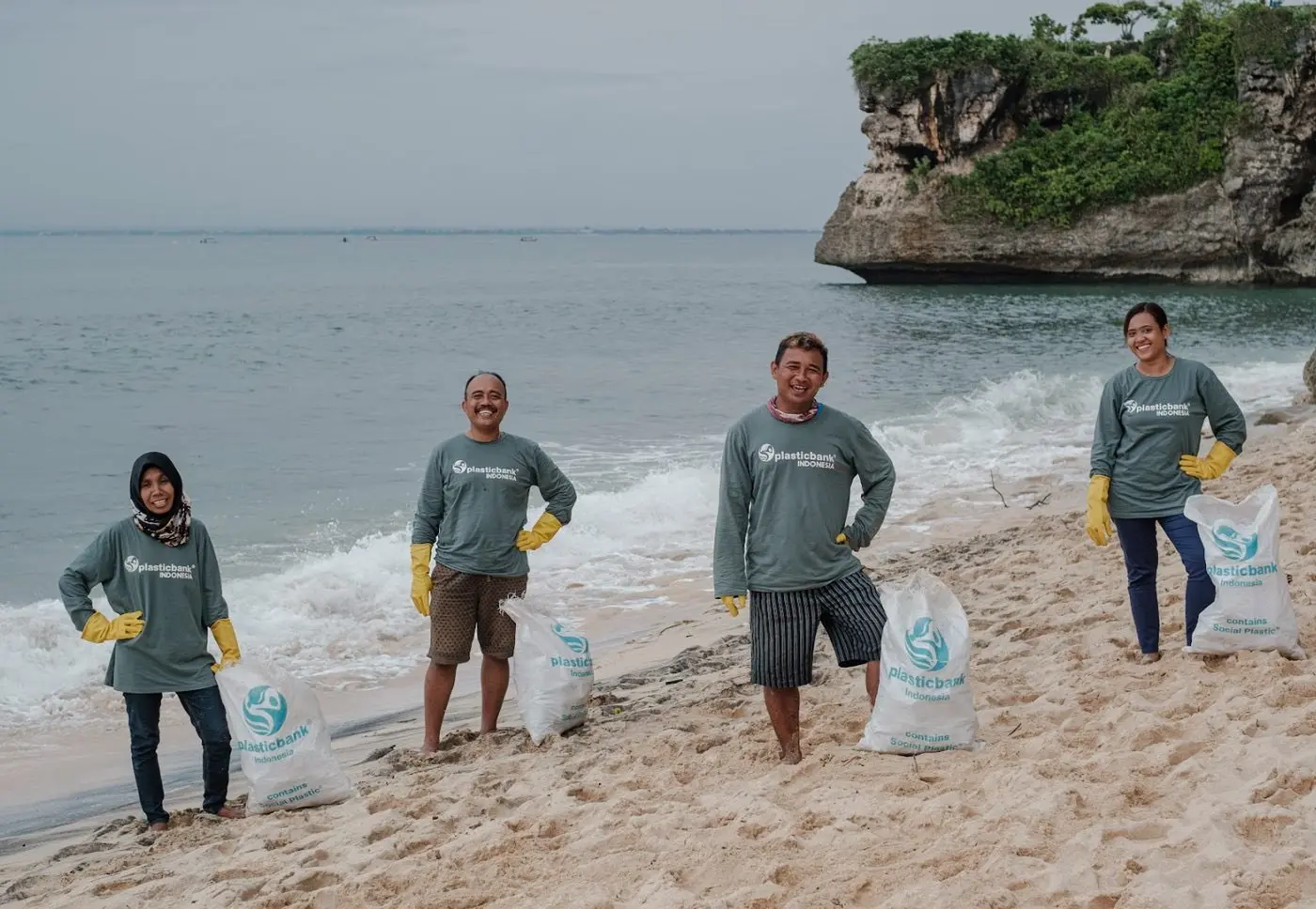 Four Plastic Bank collectors in team shirts and gloves on a beach, each with a full collection sack